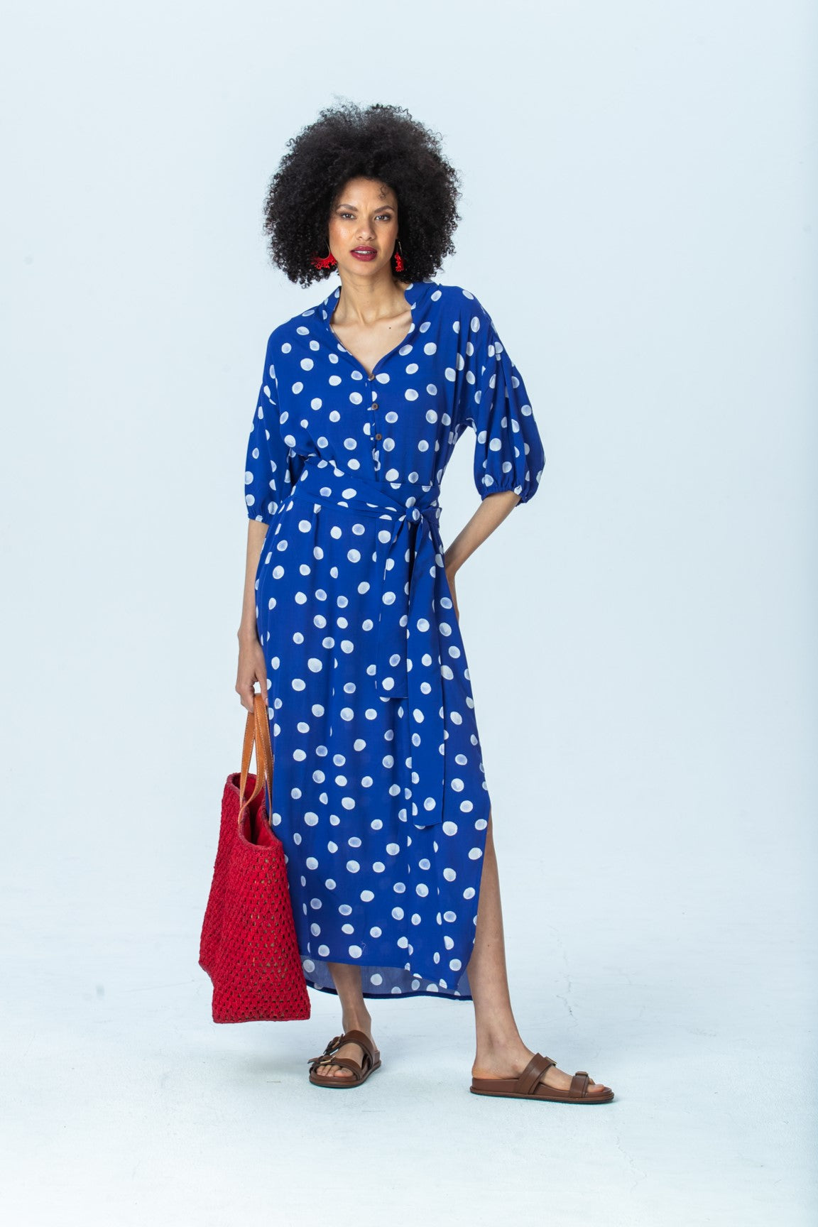 Sloane royal blue polka dot dress, front view full length, belted, holding a red woven tote