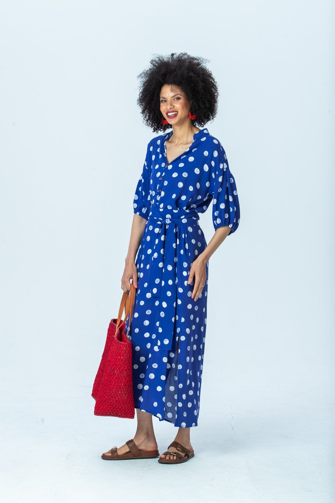 Sloane royal blue polka dot dress, three-quarter view full length, belted, holding a red tote
