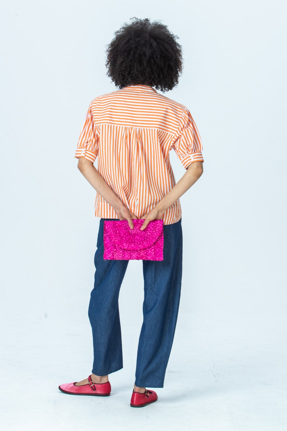 Tatum orange stripe shirt, back view full length holding a clutch behind her back